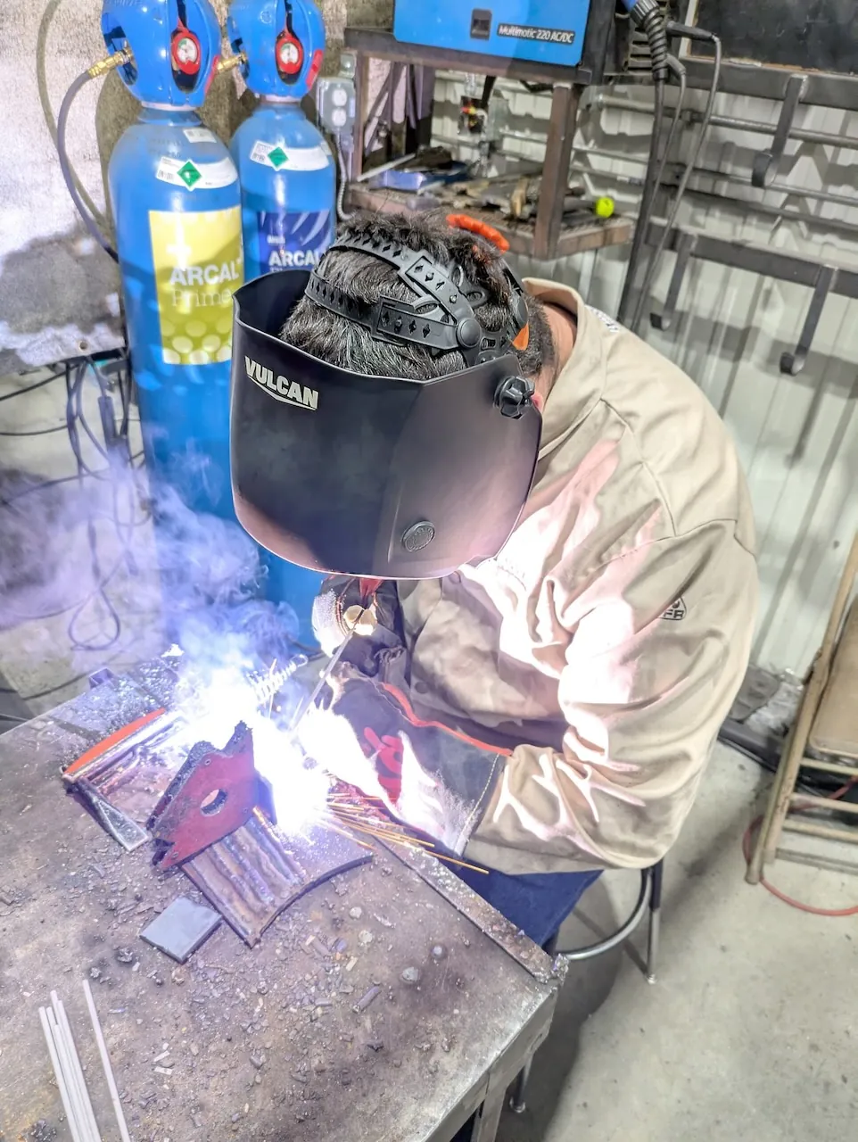 An adult welder wearing a protective helmet, jacket and gloves while welding a metal project at Kilroy’s Workshop.